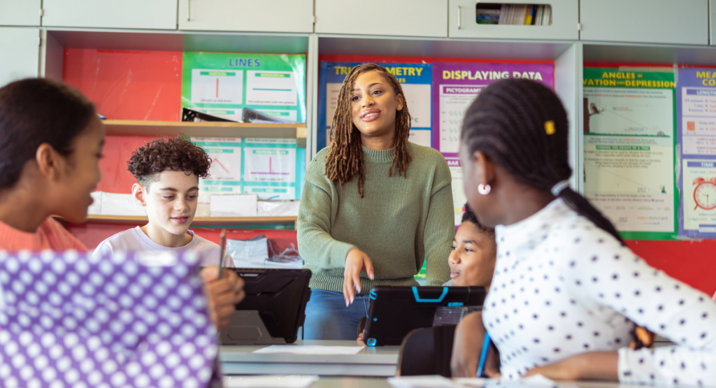 A teacher stands in the center of classroom engaging a group of students in conversation