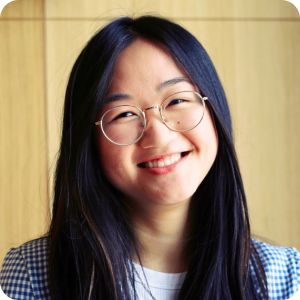 Asian woman with long black hair and round glasses smiling wearing a blue checkered blazer