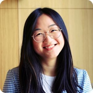 Asian woman with long black hair and round glasses smiling wearing a blue checkered blazer