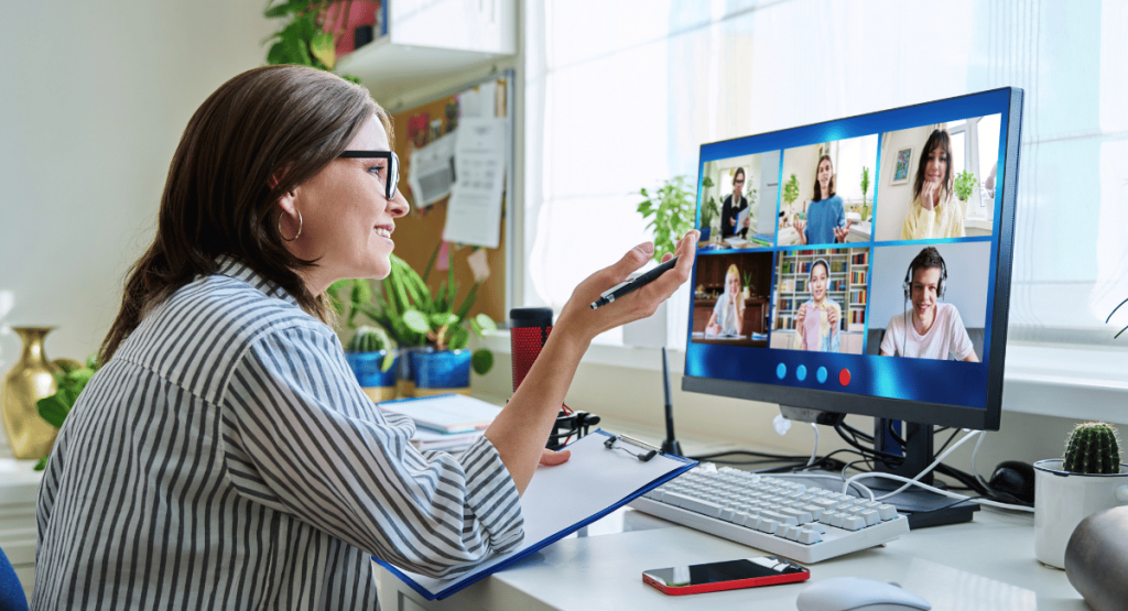 teacher with clipboard sitting in front of computer monitor with kids' faces in a virtual conference